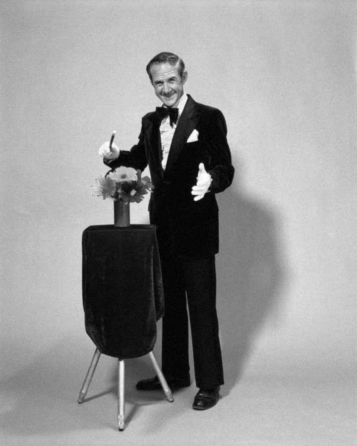 A stage magician in a tuxedo presenting a vase of flowers.
